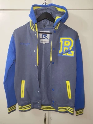 Blå och grå hoodie från R-NEAL - Snygg hoodie i collegemodell från R-NEAL med grå kropp och blå ärmar. Stora gula detaljer på fickor, muddar och huva samt ett stort gult 'R' på bröstet. Jackan har tryckknappar och broderade detaljer. Perfekt för en sportig och avslappnad stil.
