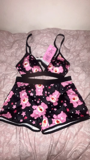 Care Bears set med shorts och topp - Sött underklädesset från Pink Soda med Care Bears-motiv. Setet består av en svart topp med smala axelband och matchande shorts, båda med rosa nallebjörnar, stjärnor och hjärtan. Materialet är mjukt och stretchigt, perfekt för en mysig känsla.