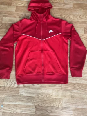 Röd Nike kofta med dragkedja - Snygg röd hoodie från Nike med vit logga på bröstet och vit detalj längs bröstet för ett billigt pris. Hoodien har dragkedja framtill, två stora fickor och justerbar huva med snören. Tillverkad i mjukt material som känns skönt mot huden. Perfekt för en sportig och avslappnad stil.