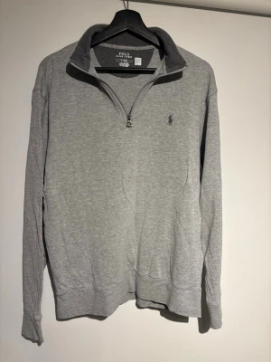 Grå halfzip från Ralph Lauren - Snygg grå långärmad half zip tröja från Polo Ralph Lauren med broderad logga på bröstet. Tröjan har hög krage och är tillverkad bomullsmaterial. Perfekt för en avslappnad och stilren look. Inför vår och sommar!
