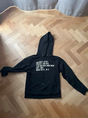 Svart hoodie från Helmut Lang - Svart hoodie från Helmut Lang med vit text på bröstet. Texten lyder 'Finest Sweats for Women and Men Est. 1986 New York, N.Y.'. Hoodien har dragsko i huvan och en klassisk magficka. Perfekt för en avslappnad och trendig stil.