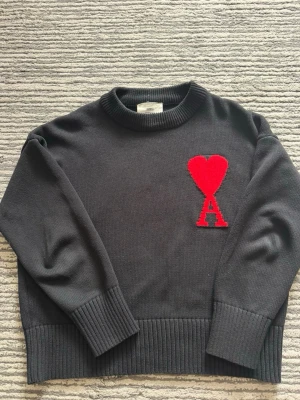 Svart stickad tröja från Ami Paris - Snygg svart stickad tröja från Ami Paris med röd hjärtlogga och bokstaven A på bröstet. Tröjan har rund hals, ribbade muddar och nederkant. Perfekt för dig som gillar stilrena plagg med en twist.