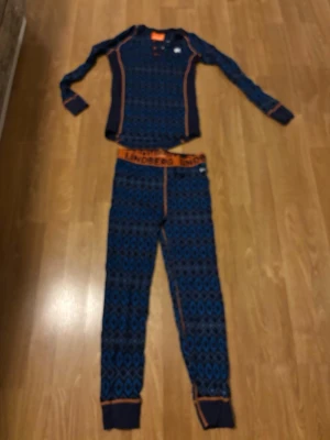 Blått underställ från Lindberg Merino ull stl 140 - Snyggt tvådelat underställ från Lindberg i blått och mörkblått med orange detaljer och grafiskt mönster. Tröjan har långa ärmar och byxorna har resår i midjan med Lindberg-tryck. Perfekt för kalla dagar och vintersport. Understället har några små hål se bilder. Merinoull, passar 140 så litet i storlek.