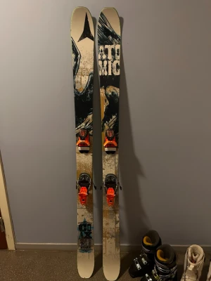 Atomic Coax twintip skidor - Säljer ett par Atomic Coax skidor med coolt grafiskt mönster i svart, beige och turkos. Skidorna har tydlig Atomic-logga och är utrustade med orangea bindningar. Perfekta för dig som gillar att åka i både pist och offpist. Skidorna är väl använda. Behöver lite kärlek för att återfå sin forna glans. 