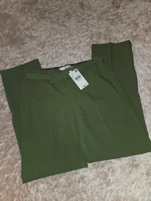Olivgröna kostymbyxor från Mango - Snygga olivgröna kostymbyxor från Mango med raka ben och hög midja. Byxorna har bälteshällor, knapp och dragkedja framtill samt diskreta pressveck. Perfekta för en clean och stilren look.