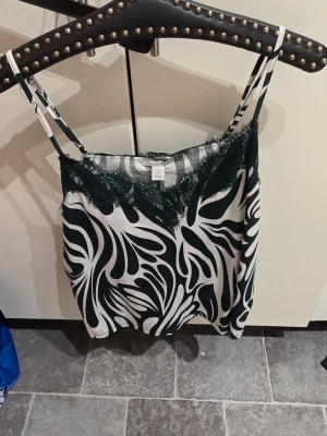 Mönstrat linne med spets från Amisu - Snyggt linne från Amisu med svartvitt abstrakt mönster och tunna axelband. Toppen har mörkgrön spetsdetalj vid urringningen som ger en extra touch. Perfekt för dig som gillar trendiga och unika plagg. Materialet känns mjukt och följsamt.