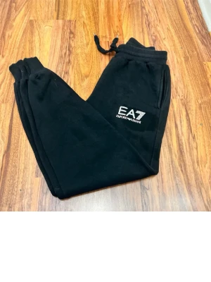 Svarta EA7 mjukisbyxor med logga - Svarta mjukisbyxor från EA7 Emporio Armani med vit logga på vänster ben. Byxorna har dragsko i midjan, muddar vid bensluten och sidofickor. Perfekta för en avslappnad och sportig stil.