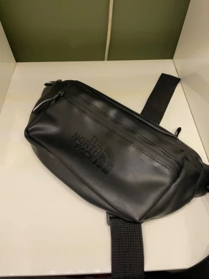 Svart shoppingväska från The North Face - Snygg svart shoppingväska från The North Face i slitstarkt material med justerbar axelrem. Väskan har flera dragkedjefack och en stilren logga framtill. Perfekt för dig som vill ha en praktisk och trendig väska till vardags.