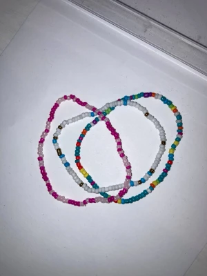 Färgglada pärlarmband i set om tre - Tre elastiska armband med små pärlor i olika färger som rosa, vitt, blått, gult och rött. Perfekta för att ge en lekfull touch till din stil. Armbanden är tunna och flexibla, gjorda av plastpärlor.