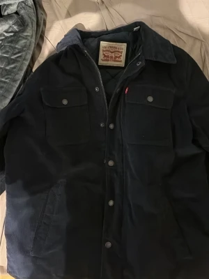 Mörk blå Levi's Jacka - Snygg svart Jacka från Levi's i mjuk manchester. Två bröstfickor med knappar, klassisk krage och knappstängning framtill. Perfekt lager-på-lager-plagg med quiltat innerfoder för extra komfort. Diskret röd Levi's-tag på bröstfickan.