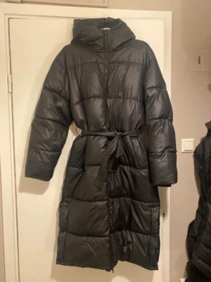 Svart lång dunjacka med bälte - Snygg svart lång dunjacka med huva och bälte i midjan. Jackan har quiltad design, dragkedja framtill och tryckknappar nertill. Perfekt för kalla vinterdagar och ger en cool och stilren look.