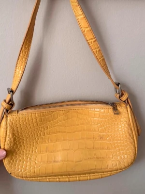 Gul axelväska i krokopräglat skinn - Snygg gul axelväska med krokodilpräglat mönster och justerbar rem. Väskan har en avlång form och stängs med dragkedja upptill. Perfekt för att addera färg till din outfit.