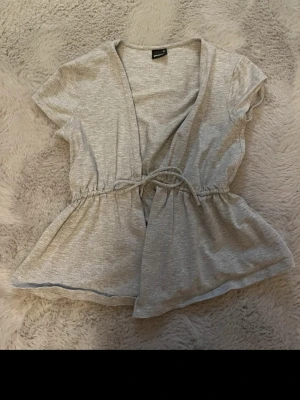 Grå omlottopp med knyt från Gina Tricot - Grå omlottopp från Gina Tricot med korta ärmar och knyt i midjan. Toppen har en v-ringad design och är lätt utsvängd nedtill för en avslappnad look. Perfekt att styla med jeans eller kjol.