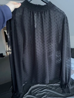 Svart transparent blus med struktur - Svart blus i transparent tyg med små strukturprickar, hög smockad halsringning och långa ärmar med smockade muddar. Blusen har knapp i nacken och ett luftigt, rakt fall. Perfekt för en trendig och stilren look.