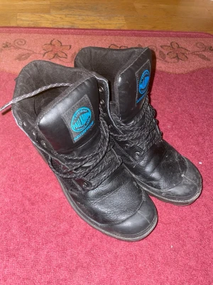 Svarta kängor från Palladium - Säljer ett par svarta kängor från Palladium med grov sula och snörning. Kängorna har både läder- och textildetaljer samt tydlig logga på sidan och plösen. Perfekta för dig som vill ha en cool och tålig look.