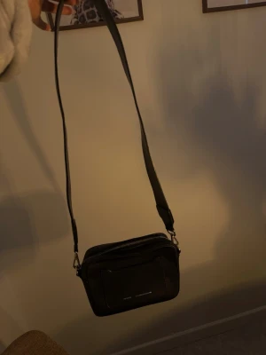 Svart axelväska från Don Donna - Snygg svart axelväska från Don Donna med justerbar rem och stilren design. Väskan har en praktisk ytterficka och detaljer i silver. Tillverkad i konstläder med präglad text på framsidan: 'Don Donna Est. 1968 Stockholm'. Perfekt för att bära det viktigaste.