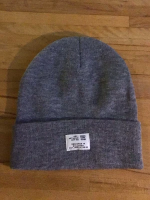 Grå ribbstickad mössa med patch - Snygg grå ribbstickad mössa med uppvikt kant och vit patch framtill med svart text. Klassisk och enkel design som passar till det mesta. Tillverkad i mjukt och stretchigt material för skön passform.