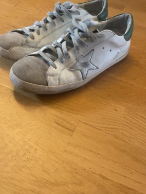Golden Goose vita sneakers med stjärna - Snygga vita sneakers från Golden Goose med grå mockadetaljer på tån och silverfärgad stjärna på sidan. Skorna har gröna hälpartier och grå skosnören. Klassisk låg modell i läder och mocka, perfekt för en trendig streetstyle-look.