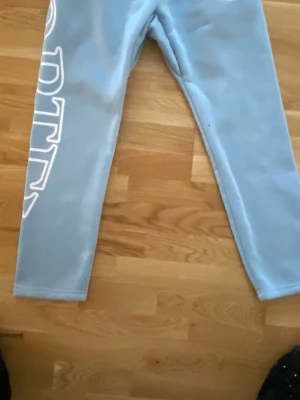 Ljusblå mjukisbyxor corteiz  - Säljer ett par ljusblå mjukisbyxor med stort vitt tryck längs ena benet och coolt grafiskt motiv. Byxorna har rak passform och resår i midjan. Perfekta för chill dagar eller när du vill ha en avslappnad stil.