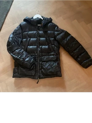 Svart dunjacka från Parajumpers - Snygg svart dunjacka från Parajumpers med huva och dragkedja framtill. Jackan har två stora fickor och ett broderat märke på ärmen. Perfekt för kalla vinterdagar och riktigt varm tack vare dunfyllningen.