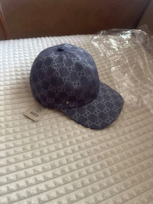 gucc* keps från  - Snygg blå keps från Gucc* med det klassiska -monogrammet över hela kepsen. Kepsen har böjd skärm och är tillverkad i ett tåligt tygmaterial. Perfekt accessoar för att lyfta din streetwear-look. 