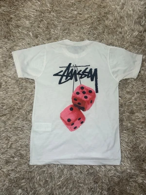 Vit Stüssy t-shirt med tärningsmotiv - Vit t-shirt från Stüssy med svart logga på bröstet och stort tryck på ryggen med två rosa tärningar och Stüssy-loggan ovanför. Klassisk passform och rund halsringning. Perfekt för dig som gillar streetwear och vill sticka ut med en unik design.