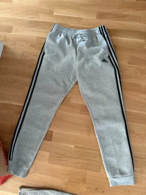 Gråa Adidas mjukisbyxor med svarta ränder - Gråa mjukisbyxor från Adidas med klassiska svarta ränder längs sidorna och Adidas-logga på vänster lår. Byxorna har resår i midjan med dragsko och muddar vid bensluten. Perfekta för en avslappnad och sportig stil.