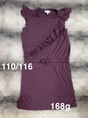 Lila klänning med volang och knytband - Söt lila klänning från pompdelux med volangdetalj snett över framsidan och små volanger vid axlarna. Klänningen har ett knytband i midjan och är ärmlös, perfekt för varma dagar.🌸Fint använt skick. Storlek 110/116 