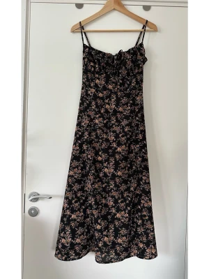 Svart blommig långklänning - Svart långklänning med tunna axelband och blommigt mönster i beige och rosa toner. Klänningen har en söt rosettdetalj framtill och smickrande passform med lätt utställd kjol. Perfekt för varma dagar och sommarens alla tillfällen. Mycket gott skick, som ny. 