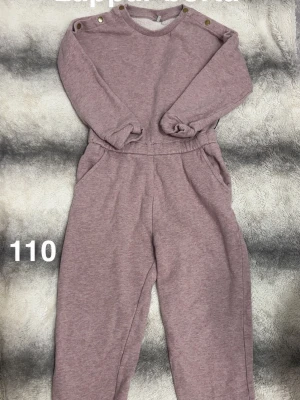 Ljuslila jumpsuit med knappar - Mysig ljuslila jumpsuit i mjukt sweatshirtmaterial med långa ärmar och ben. Rund halsringning med dekorativa guldknappar på axlarna och praktiska fickor i sidorna. Perfekt för en avslappnad och chill stil.🌸 Fint använt skick. Antydan till väldigt svag fläck på knät. Lappar är borta. Storlek 110