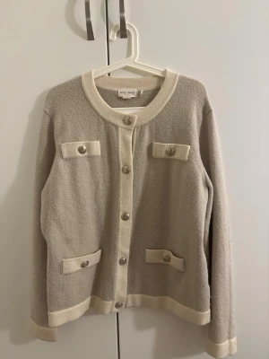 Beige kofta från Holly & Whyte - Stilren beige kofta från Holly & Whyte med kontrasterande ljusa kanter och dekorativa fickor framtill. Koftan har rund halsringning, långa ärmar och knappar i silverfärg. Perfekt för en klassisk och chic look.