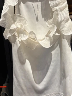 Vit blus med volang från Misty mynk - Supersnygg vit blus med stora, plisserade volanger längs urringningen. Tillverkad i ett ribbat tyg som ger en trendig känsla. Perfekt för dig som vill sticka ut med en unik och feminin look.