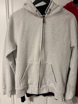 Carhartt hoodie  - Ljusgrå hoodie med dragkedja från Gul & Blå. Klassisk modell med huva och dragsko, samt två stora fickor framtill. Mjuk bomullsblandning och ribbade muddar vid ärmslut och nederkant. Liten gul logga på ärmen för en extra detalj.
