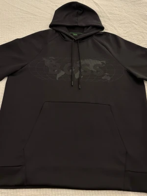 Svart Hugo Boss Soody Diamond hoodie  - Hugo Boss Soody Diamond hoodie i svart färg.  Stilren huvtröja från BOSS Menswear med normal passform. Modellen har ett diskret grafiskt klotmotiv samt en BOSS-logotyp med strassprydda detaljer som ger ett exklusivt men nedtonat uttryck. Tillverkad i stretchinterlock med mjuk känsla.  Huva med bomullsfoder för extra komfort, långa ärmar och klassisk kangaroo-ficka framtill. Muddar i ärmslut och nederkant.  Äkta Hugo Boss i mycket bra skick – endast använd några få gånger, inga fläckar eller hål.