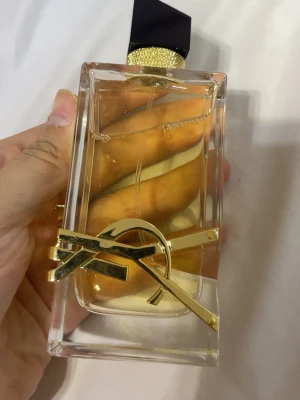 Yves Saint Laurent Libre Eau de Parfum - Snygg parfymflaska från Yves Saint Laurent, modell Libre. Flaskan är rektangulär i genomskinligt glas med guldig detalj och svart asymmetrisk kork. Innehållet är ljusgult och flaskan har en lyxig känsla med YSL-logga i metall framtill.