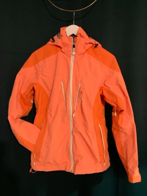 Orange vindjacka från Tenson - Snygg orange vindjacka från Tenson med dragkedja framtill, flera praktiska fickor och justerbar huva. Jackan har sportig design med ljusare och mörkare orange partier samt detaljer i vitt. Perfekt för blåsiga dagar och outdoor-aktiviteter.