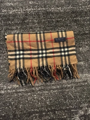 Beige rutig halsduk från Burberrys - Klassisk beige halsduk från Burberrys med ikoniskt rutmönster i svart, vitt och rött. Tillverkad i 100% lammull och har fransar i båda ändar. Perfekt accessoar för att lyfta din outfit med en tidlös brittisk stil.