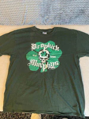 Grön Dropkick Murphys t-shirt - Cool grön t-shirt från Gildan med stort Dropkick Murphys-tryck på bröstet. Trycket har en dödskalle och fyrklöver i grönt och vitt. Klassisk passform och rund hals. Tillverkad i kraftig bomull som känns rejäl och bekväm. Storlek M/L