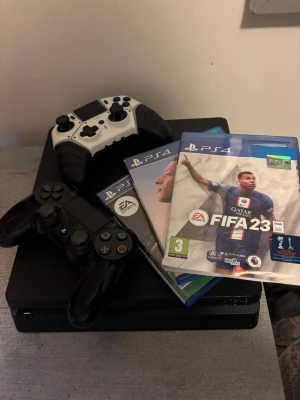 PlayStation 4 med två handkontroller - Säljer en svart PlayStation 4 med fyra handkontroller, två svarta och två vit/svarta. Medföljer flera PS4-spel, bland annat FIFA 23. Perfekt för gamingkvällar med kompisar. Konsolen har en stilren, rektangulär design i plast.