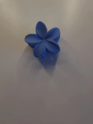 Blå blomformad hårklämma - Söt hårklämma i blått med blomformad design. Klämman är tillverkad i plast och har fem kronblad som ger ett lekfullt och trendigt intryck. Perfekt för att snabbt sätta upp håret med en färgklick och en rolig detalj.