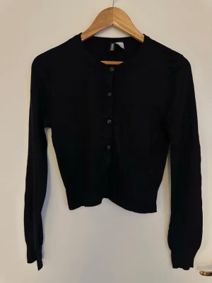 Svart cropped cardigan från H&M - Svart långärmad cardigan från H&M med rund halsringning och knappar framtill. Modellen är cropped och har en enkel, stilren design som passar till många olika outfits.