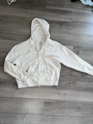 Ljusgrå zip hoodie med huva - Ljusgrå hoodie från Gildan med dragkedja framtill och två fickor. Klassisk modell med huva och dragsnören. Tillverkad i mjukt sweatshirtmaterial, perfekt för chill dagar. Liten vit brodyrdetalj på bröstet.