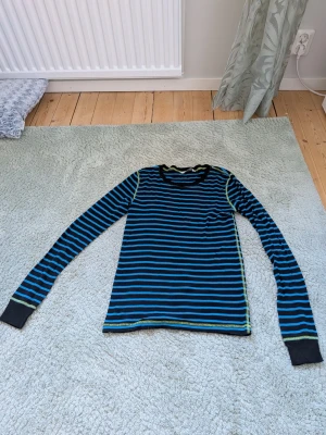 Randig långärmad underställströja blå/svart - Långärmad underställströja tröja med blå och svarta horisontella ränder samt gröna sömmar. Tröjan har rund halsringning och svarta muddar vid ärmsluten. Perfekt för träning utomhus på vintern.