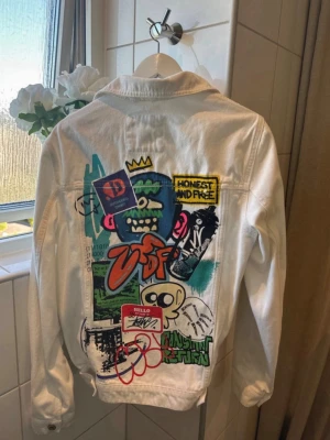 Vit jeansjacka från Zara - Vit jeansjacka från Zara med coola graffiti- och patchdetaljer på ryggen. Jackan har klassisk krage, knäppning framtill och två bröstfickor med lock. Färgglada tryck och broderier ger jackan en unik streetstyle-look.