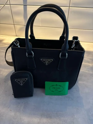 Svart Prada handväska - Snygg svart handväska från Prada i nylon med dubbla handtag och avtagbar axelrem. Väskan har silverfärgade detaljer och klassisk Prada-logga framtill. Medföljer matchande nyckelplånbok och äkthetskort. Perfekt för dig som gillar stilrena accessoarer.