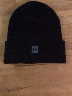 Svart mössa från Under Armour - Säljer en svart mössa från Under Armour med uppvikt kant och en liten logotyp framtill. Klassisk och enkel design som passar till det mesta. Perfekt för kalla dagar och enkel att matcha med olika outfits.