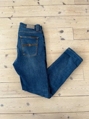 Blå jeans från nudie - Säljer ett par klassiska blå jeans från Nudie Jeans med slim passform och snygga slitningar. Jeansen har fem fickor, orange söm på bakfickan och är gjorda i mjuk denim. Perfekta för en avslappnad och trendig look. Jeansen är i mycket bra skick förutom ett igensytt hål vid skrevet. Hålet är fixat av nudie själva men det är inget man tänker på vid användning. Modell är Lean Dean och de är storlek 30W och 32L. Vid frågor eller funderingar är det bara att fråga Mvh Olof.