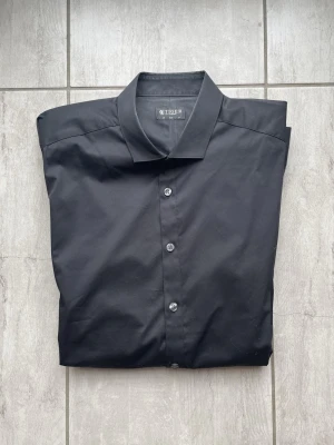 Svart skjorta från Tiger of Sweden - Klassisk svart skjorta från Tiger of Sweden med knappar framtill och lång ärm. Skjortan har en stilren design med normal krage och är tillverkad i ett slätt, tunt tyg som känns skönt mot huden. Perfekt för dig som gillar en clean och tidlös look. Vid frågor och funderingar skriv gärna!