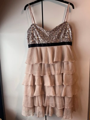 Paljettklänning med volanger - Supersöt kortklänning med glittrig paljettöverdel i silver och tunna axelband. Kjolen har flera lager volanger i ljus beige ton och ett svart band markerar midjan. Perfekt för dig som vill sticka ut med en unik look.