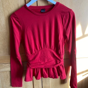 Röd peplumtopp från Gina Tricot - Snygg röd topp från Gina Tricot med lång ärm och markerad midja och peplum. Toppen har volang nedtill och en figurnära passform som framhäver siluetten. Perfekt för dig som vill ha en färgstark och trendig look. Använd en gång.